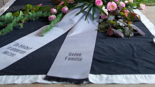 Blumenarrangement mit rosa Rosen und eine Trauerschleife vor einem Kreuz in einer Kapelle.