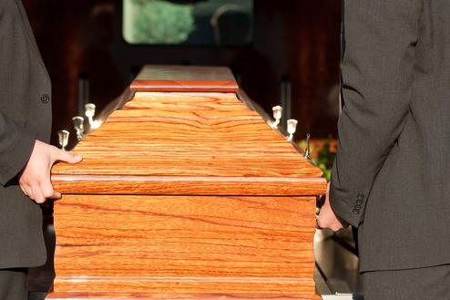 Zwei Trauergäste in Anzügen tragen einen eleganten, braunen Holzsarg aus einem Fahrzeug.