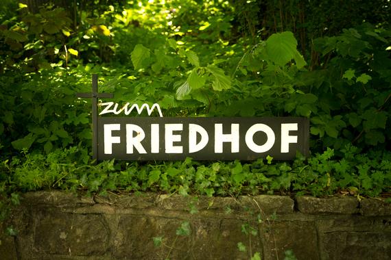 Schwarzes Schild mit der Aufschrift „zum FRIEDHOF“ und einem Kreuz, umgeben von grünem Laub.