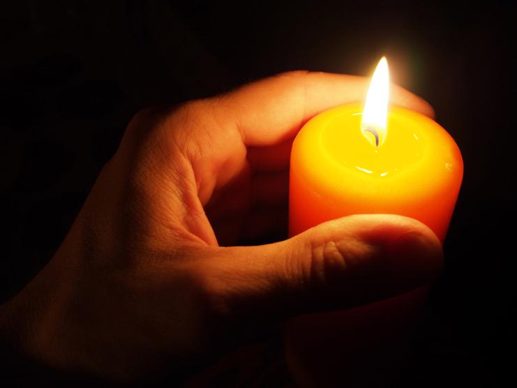 Eine Hand hält eine brennende orangefarbene Kerze in einem dunklen Umfeld.