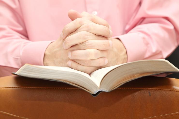 Hände eines Mannes sind gefaltet über einem offenen Buch, auf einem braunen Ledersofa. Der Mann trägt ein rosa Hemd.